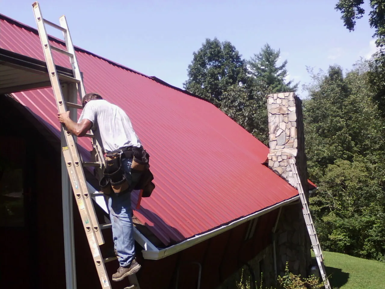 Roofer on a ladder beside a red metal roof and stone chimney — Soffit & Fascia Repair by Roofing Brothers in Peoa